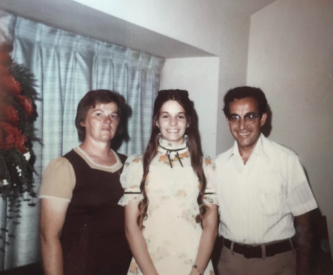 Three people stand indoors in front of a window with blue curtains. A young woman in a floral dress is between an older woman in a brown dress and an older man in a short-sleeve white shirt and glasses. Flowers are visible on the left.