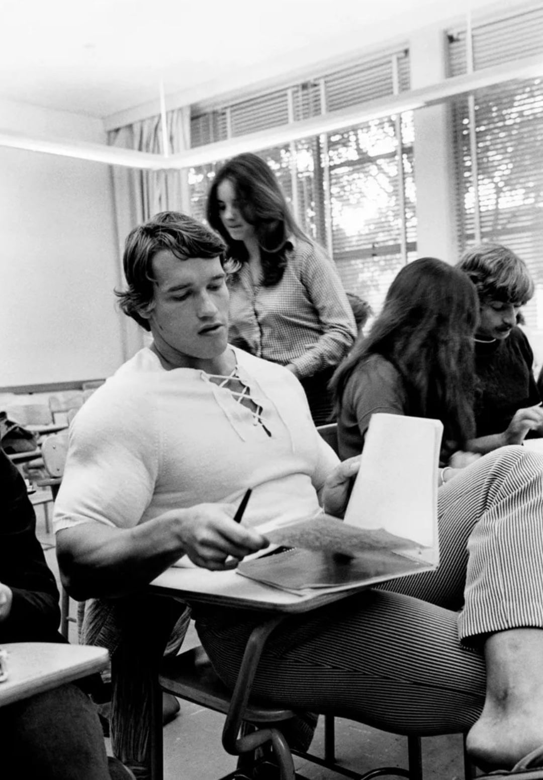 A young man with a muscular build sits at a classroom desk, writing in a notebook. Other students sit nearby, and a woman stands behind him. Large windows let in daylight, creating a bright, studious atmosphere.