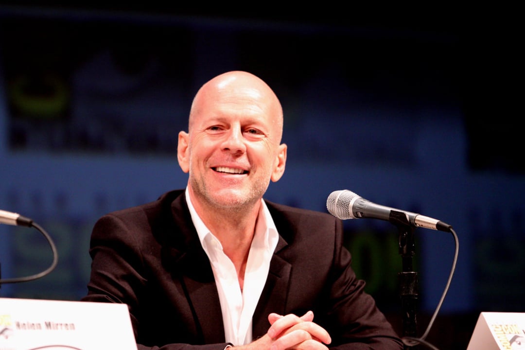 A man in a black suit and white shirt sits at a table, smiling, with a microphone in front of him. The background is blurred with text and logos.