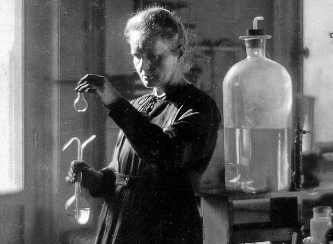 A woman in a dark dress conducts an experiment in a laboratory, carefully holding laboratory glassware. Large scientific equipment and a bottle of liquid are seen on a table beside her. The scene is in black and white.