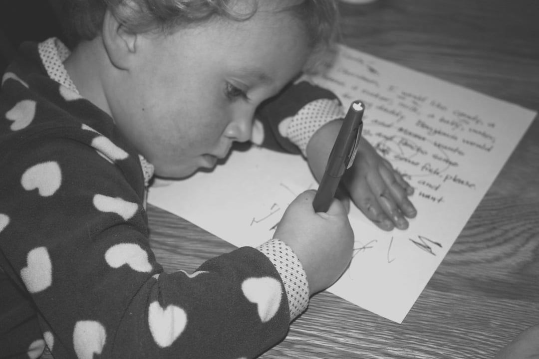 A young child in heart-patterned pajamas lies on a wooden table, holding a pen and scribbling on a piece of paper with handwritten text.