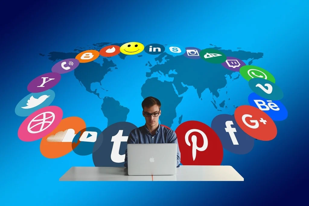 A man sits at a desk using a laptop, with various colorful social media icons arranged in an arc above him against a blue background with a world map.