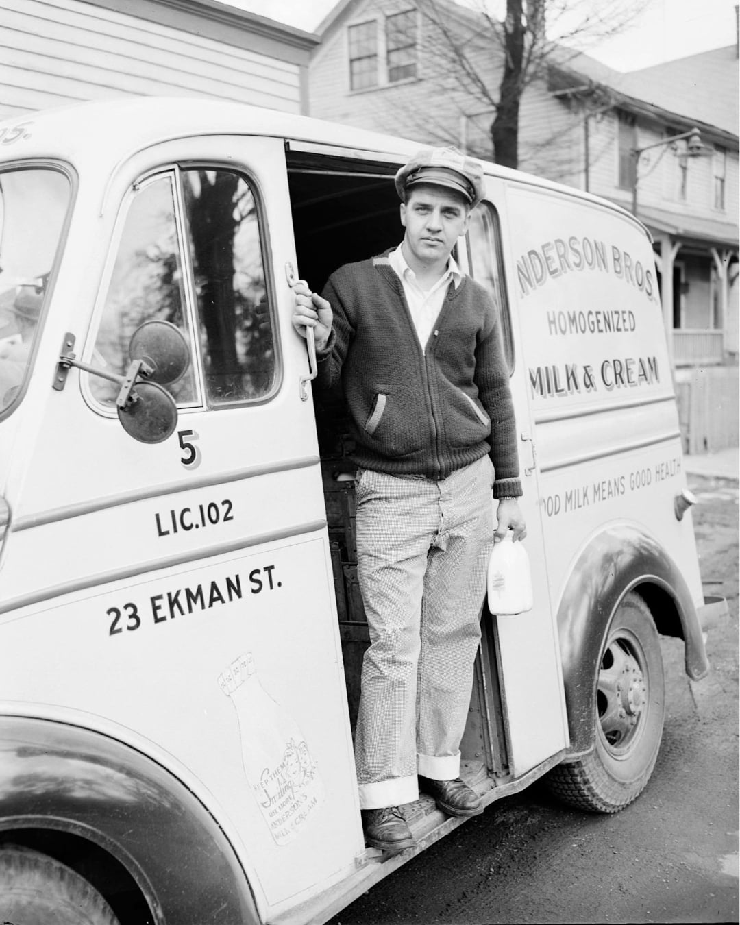 A man in a cap and cardigan stands in the open doorway of a vintage Anderson Bros milk delivery truck, holding a glass milk bottle. The truck is parked on a residential street with houses in the background.