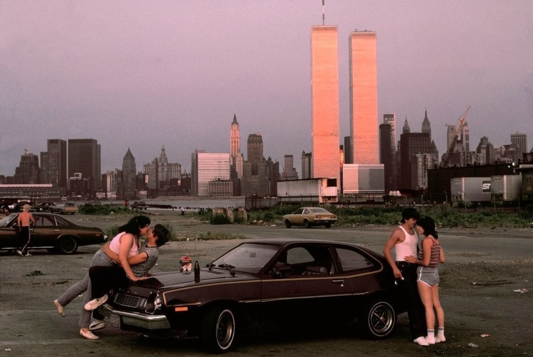 Two couples are by a dark car in a lot with the New York City skyline and Twin Towers behind them at sunset. One couple kisses on the hood while the other stands talking nearby. The city buildings are bathed in pinkish light.