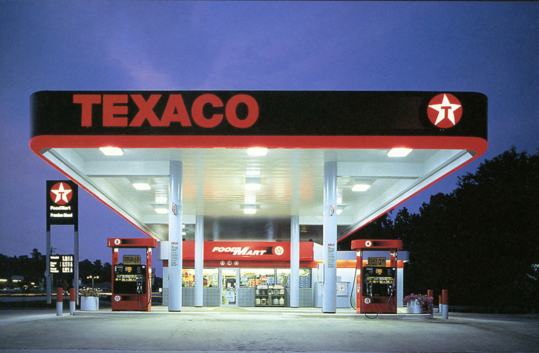 A Texaco gas station with red and white branding, two fuel pumps, and a convenience store labeled "Food Mart" at dusk, with lights illuminating the canopy and store entrance. Trees are visible in the background.