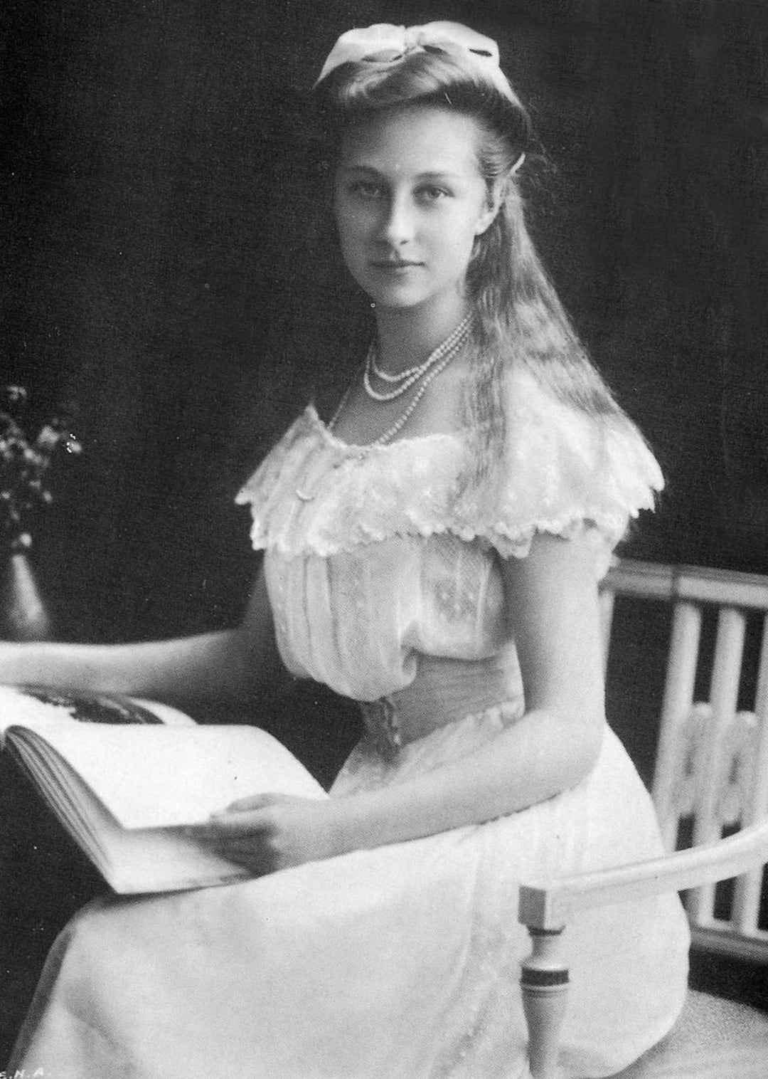 A young woman in a white, lace-trimmed dress and pearls sits holding an open book, gazing calmly at the camera. Her long hair is loose, adorned with a ribbon, and she is seated in a light-colored chair.