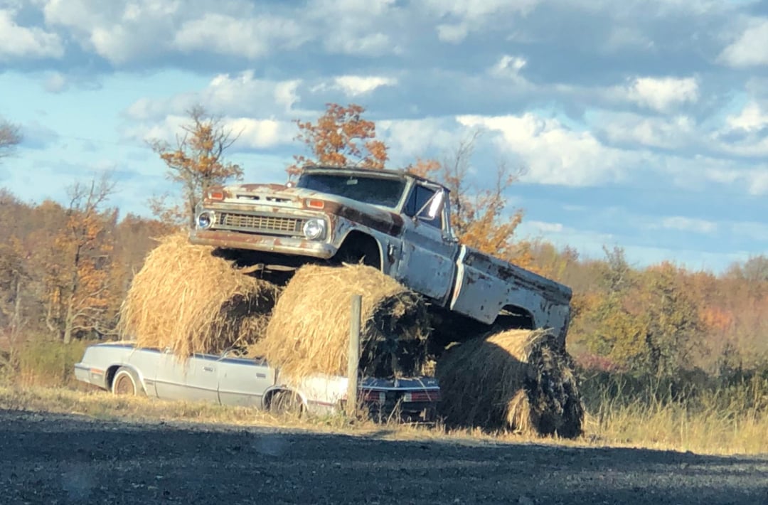 A rusty old pickup truck sits atop large bales of hay, which are stacked on top of two abandoned cars in a grassy field under a partly cloudy sky. Autumn trees are visible in the background.