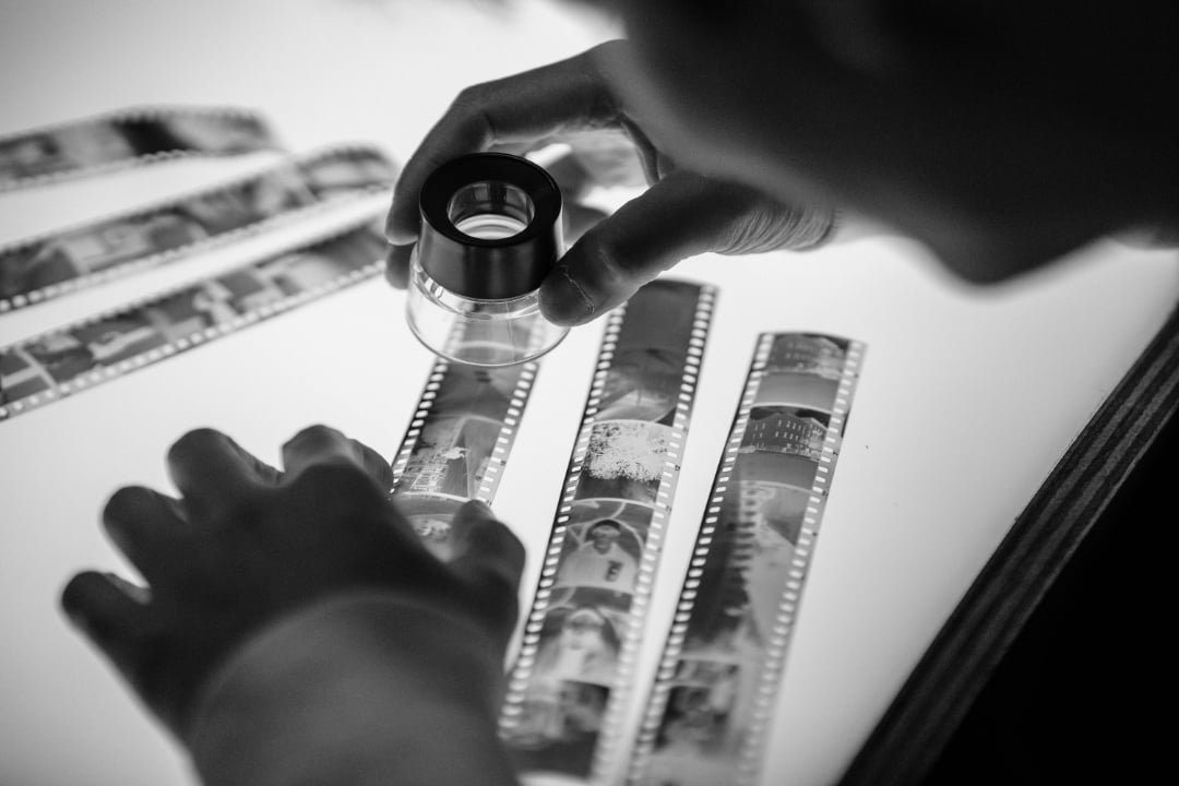 A person examines strips of photographic film negatives on a light table using a magnifying loupe, with their hands adjusting the film in a black and white image.