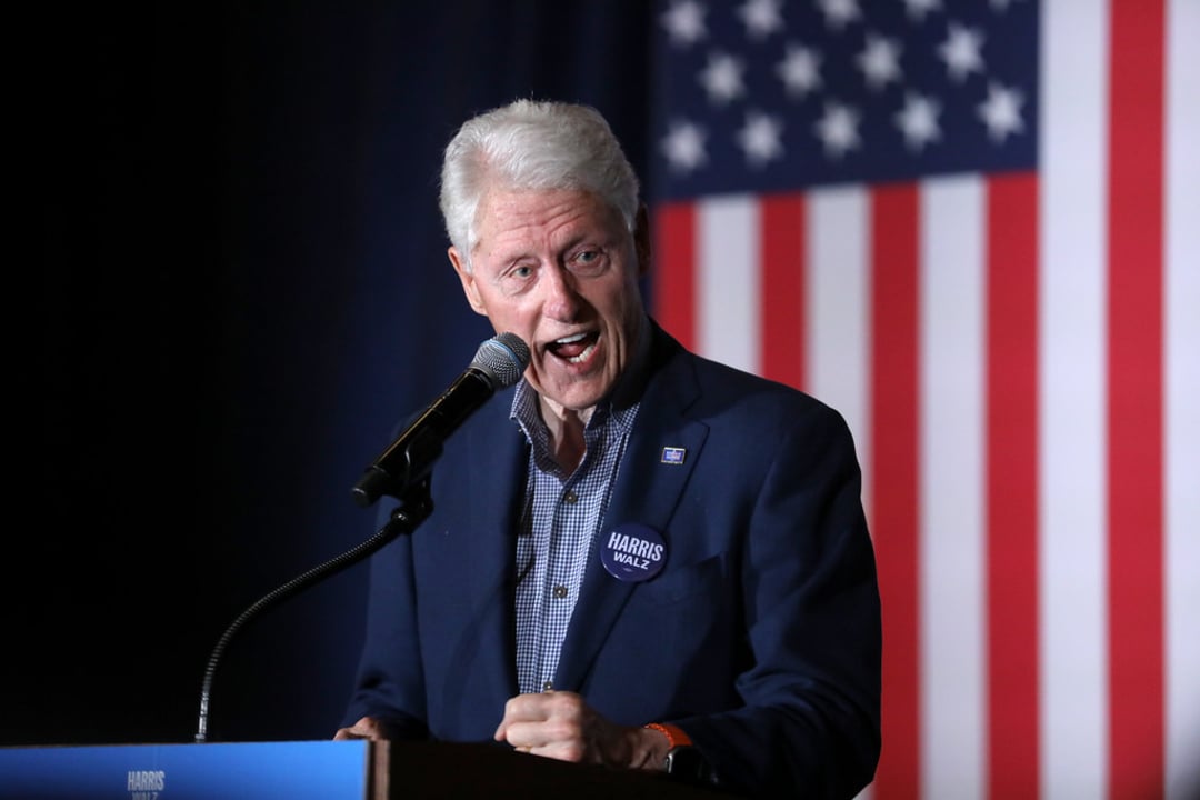 An older man with white hair speaks passionately at a microphone, wearing a dark blue blazer and a "Harris" campaign button, with a large American flag in the background.