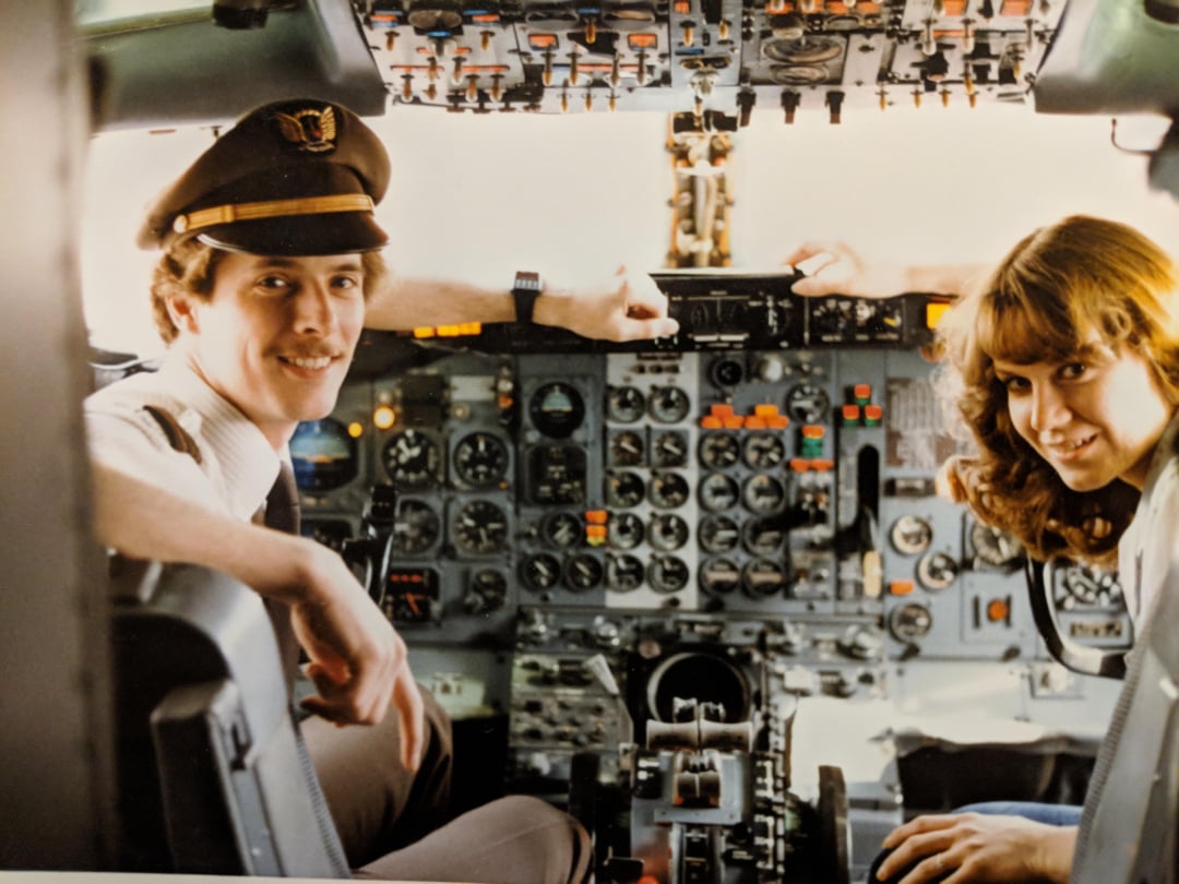 A pilot in uniform and a woman with long hair sit smiling in the cockpit of an airplane, surrounded by various control panels and instruments.