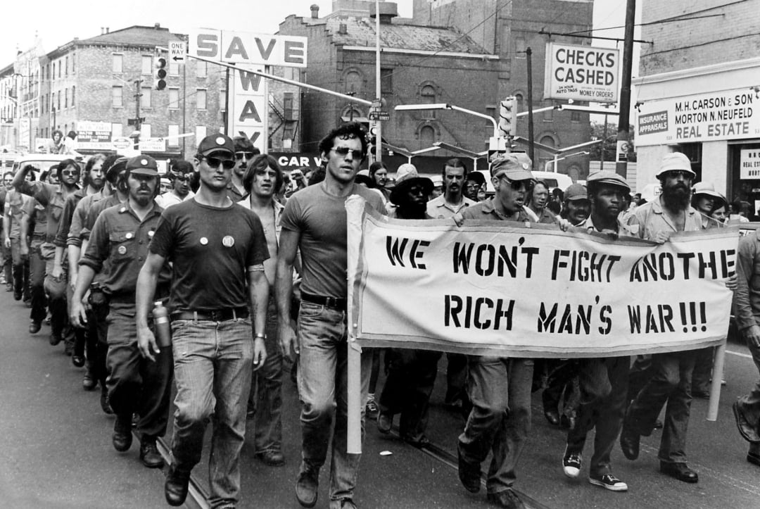 A group of demonstrators march down a city street holding a banner that reads "WE WON'T FIGHT ANOTHER RICH MAN'S WAR!!!" Other protest signs, including “SAVE WAR,” are visible in the crowd and buildings line the background.