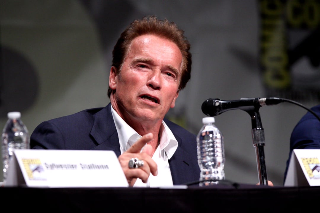 A man in a suit speaks into a microphone at a panel discussion, gesturing with his hand. Water bottles and name cards are on the table in front of him. The background features a Comic-Con logo.