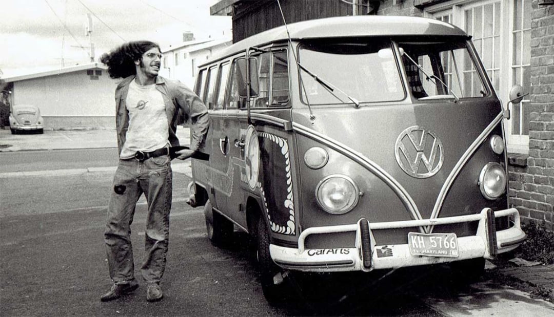 A smiling man with long curly hair stands beside a vintage Volkswagen van decorated with designs and the number 59, parked on a street near a brick building. The photo is black and white and has a retro feel.