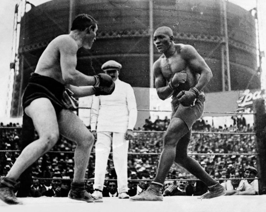 Two male boxers face each other in a boxing ring with fists raised, as a referee watches. A large crowd fills the stands and a large round building is visible in the background. The image is in black and white.