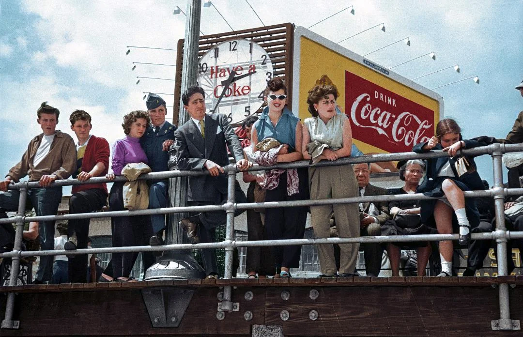 A group of young people in 1950s-style clothing stand by a metal railing on a pier beneath a large Coca-Cola sign and clock, looking out with varied expressions under a partly cloudy sky.