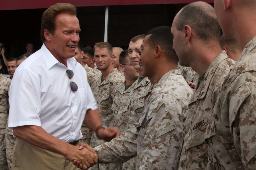 A man in a white shirt shakes hands and smiles with a group of uniformed soldiers outdoors, surrounded by others who are watching and smiling.