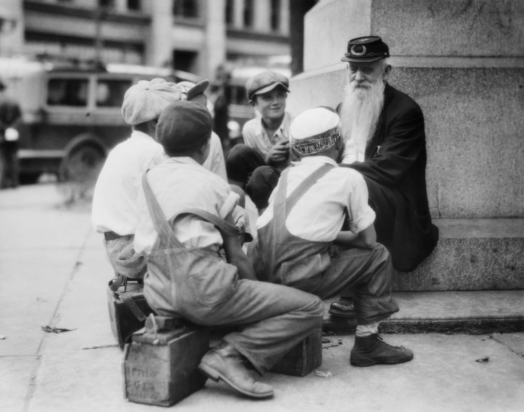 A group of boys in caps and suspenders sit on boxes around an elderly man with a long white beard and hat, who is leaning against a stone column, telling them a story on a city street.