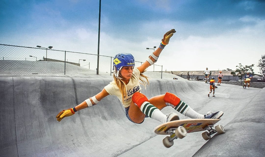 A skateboarder wearing a helmet, gloves, knee pads, and striped socks performs a trick on the edge of a concrete skate park bowl, with other skaters in the background.