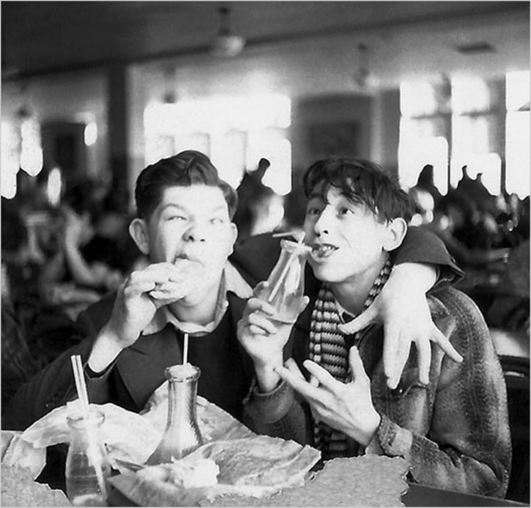 Two young men sit close together at a table, smiling while eating sandwiches and drinking from glass bottles. One has his arm around the other in a friendly gesture. The scene appears to be a lively cafeteria.