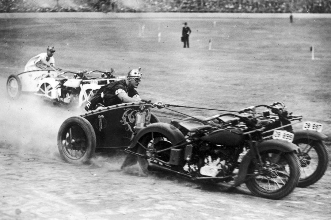 Two men in costumes race on a dirt track, each riding a motorcycle and holding reins attached to sidecars made to look like horses, kicking up dust as they speed along. Spectators can be seen in the background.