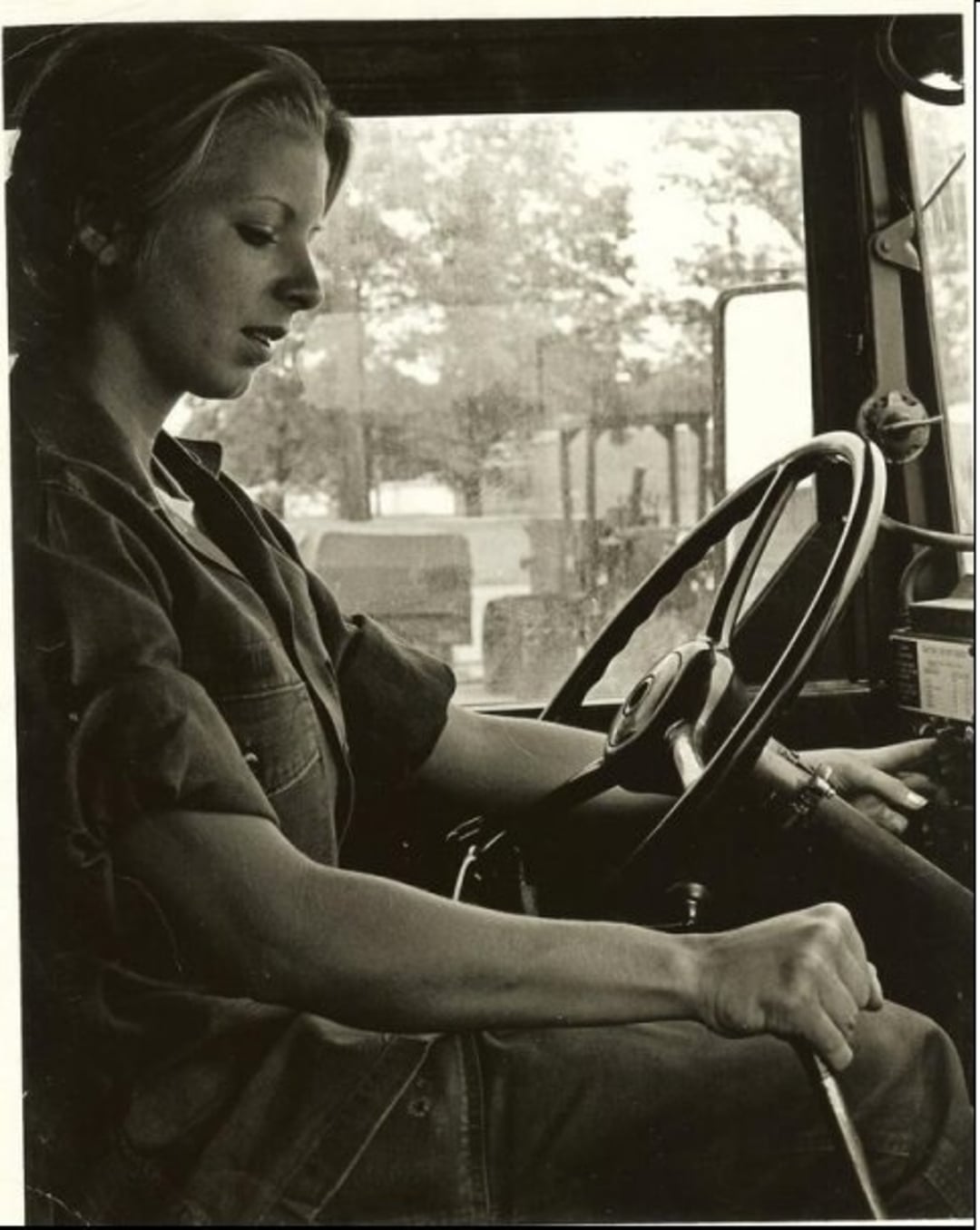 A woman in a uniform sits behind the wheel of a vehicle, possibly a truck, focused as she operates the gear shift. Sunlight filters through the window, illuminating her face and arm.