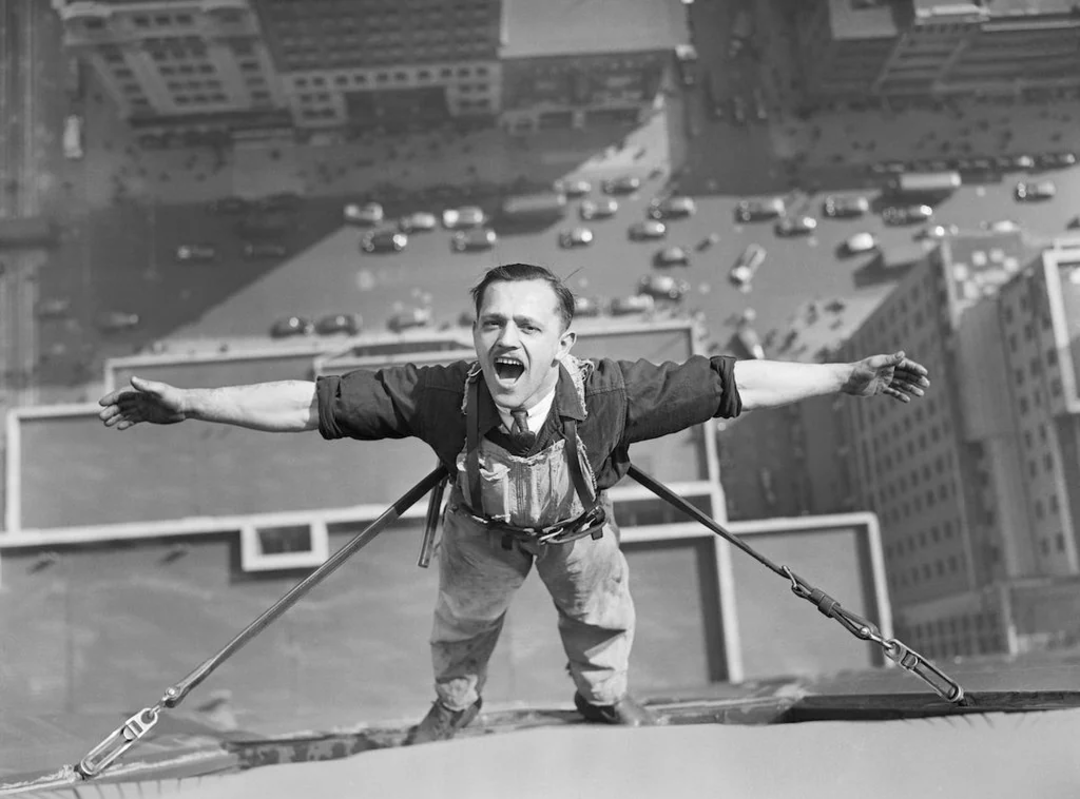 A construction worker, harnessed for safety, stands on a steel beam high above a city street, arms outstretched and mouth open, with traffic and tall buildings visible far below.