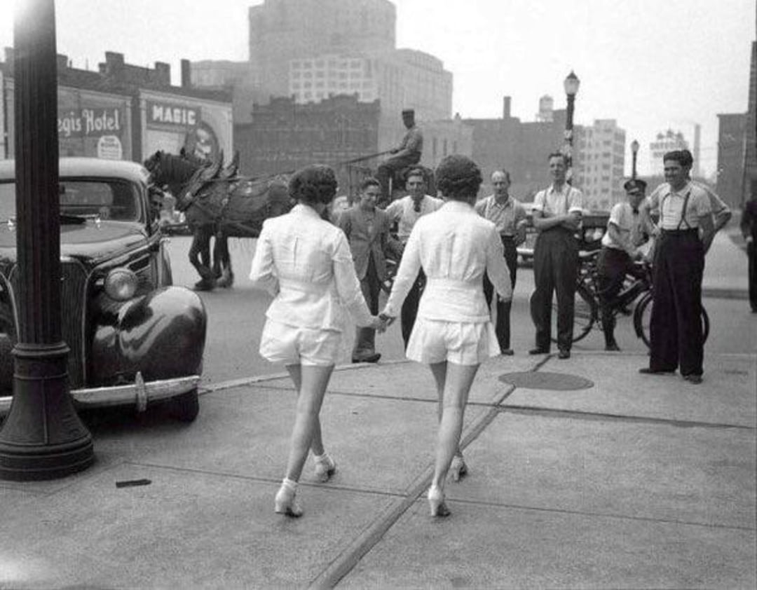 Two women in matching white outfits walk down a city sidewalk, while a group of men standing nearby watch them. A vintage car and a horse-drawn carriage are visible, suggesting the photo is from the early 20th century.