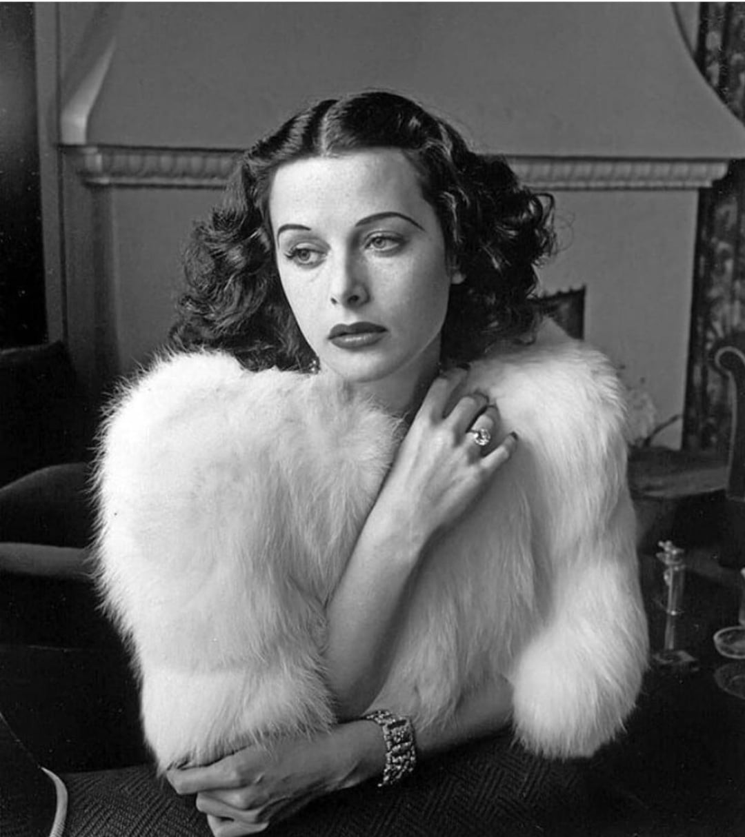 A woman with dark, wavy hair and elegant makeup gazes thoughtfully to the side while wearing a luxurious white fur stole, a sparkling ring, and a bracelet, sitting indoors in front of a mantelpiece.