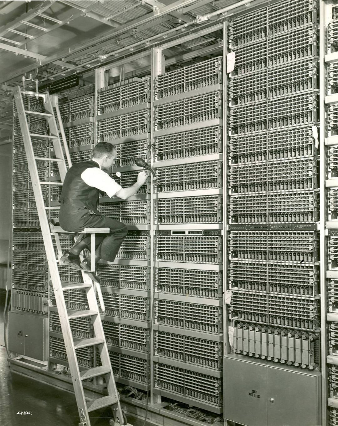 A man sits on a stepladder working with wires inside a large, complex wall of vintage electronic equipment or switchboards.