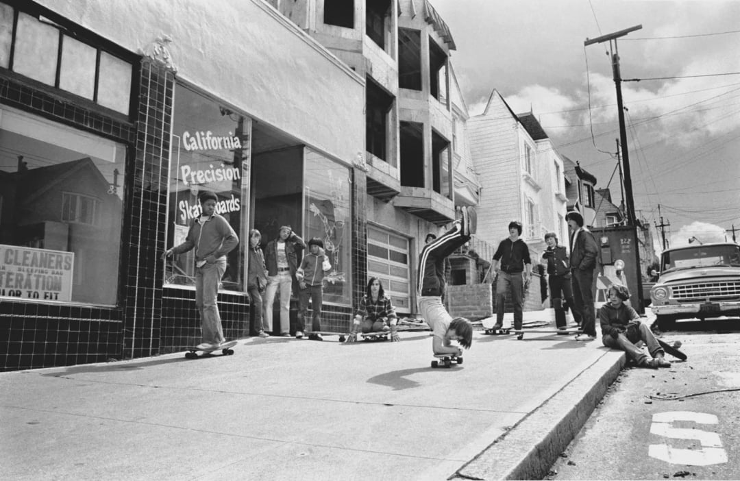 A group of kids with skateboards gather on a sloped city sidewalk. One child does a handstand on a skateboard, while others watch. An old truck is parked at the curb near a shop and partially built houses.