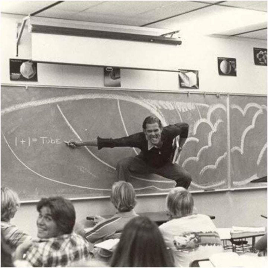 A teacher enthusiastically poses in a crouched stance on a desk in front of a chalkboard decorated with a large wave drawing, while students in the classroom smile and watch.