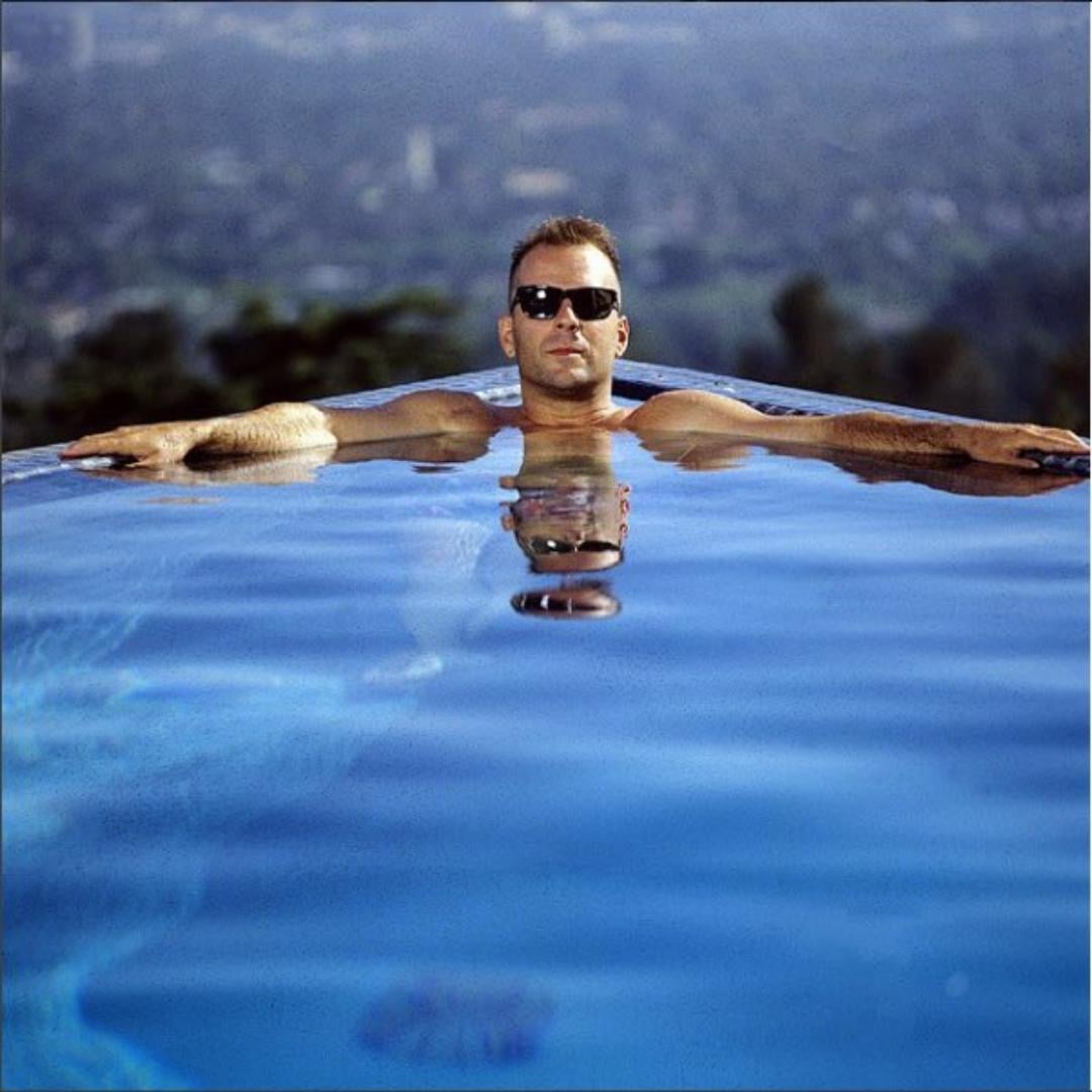 A man wearing sunglasses relaxes in an infinity pool, arms stretched out on the edge, with his reflection visible in the water and a blurred natural landscape in the background.