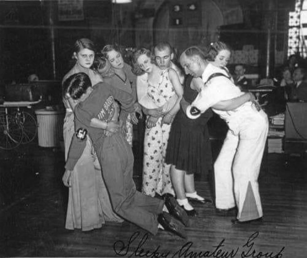 Six people, four women and two men, stand in a line indoors, appearing to play a group game. The men, in sailor and sports uniforms, lean dramatically, supported by the women, while everyone laughs or smiles.