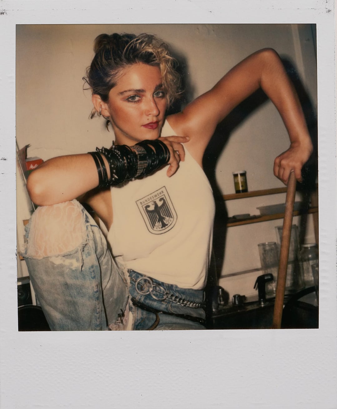 A person with short blonde hair and bold makeup poses confidently in a kitchen, wearing a sleeveless shirt with an emblem and ripped jeans, accessorized with multiple black bracelets on one arm.