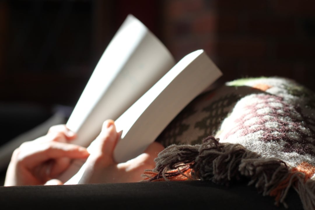 A person lies under a patterned blanket, holding and reading an open book. Sunlight softly illuminates their hands and the book, creating a cozy and peaceful atmosphere.