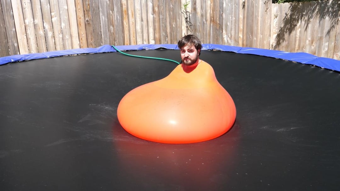A man sits inside a large, partially inflated orange balloon on a black trampoline in a fenced backyard, with only his head visible outside the balloon.