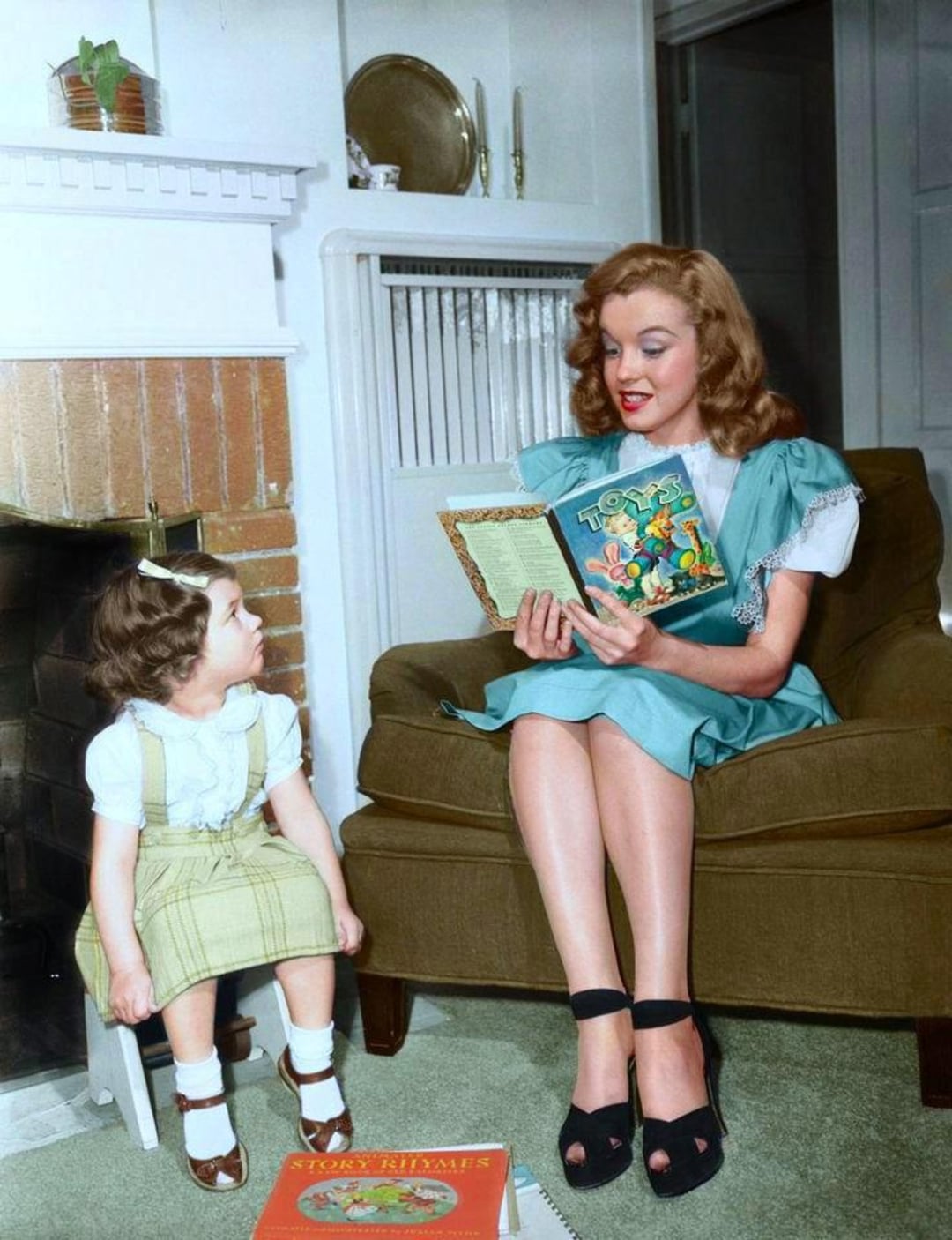 A woman in a blue dress sits in an armchair reading a storybook to a young girl in a plaid dress. They are in a cozy living room with a fireplace, radiator, and children's books on the floor.