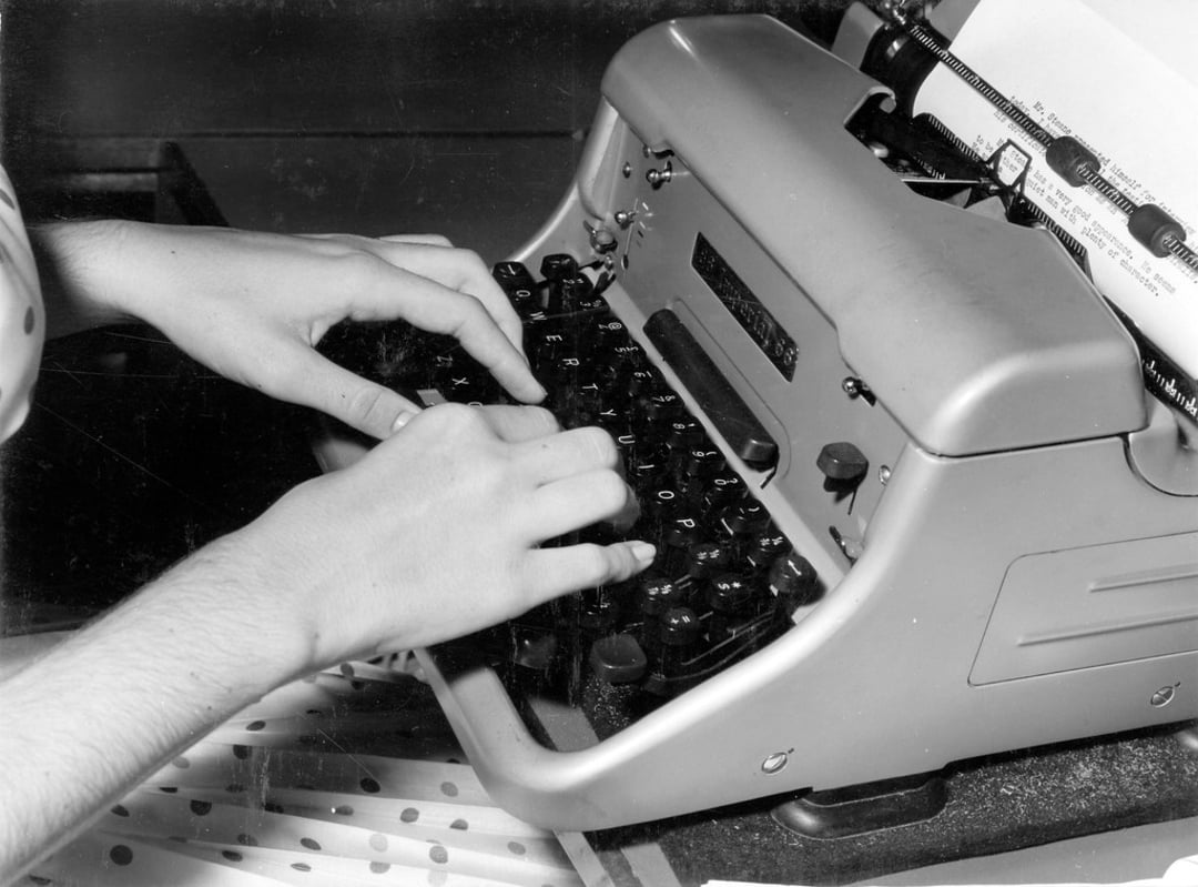Black and white photo of a person’s hands typing on a vintage typewriter, with a sheet of paper inserted and some papers with holes visible nearby.