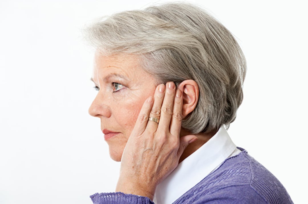 An older woman with short gray hair touches her ear with both hands, appearing concerned. She is wearing a white collared shirt and a purple sweater, shown in a side profile against a plain background.