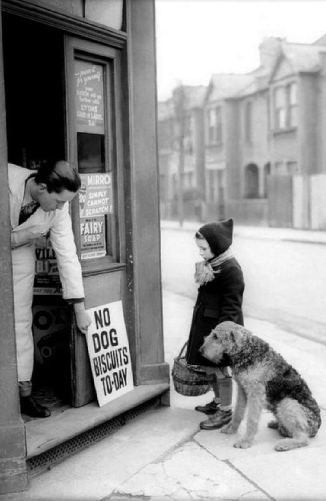 A man points to a sign reading "NO DOG BISCUITS TO-DAY" at a shop entrance, while a young girl holding a basket and a dog on a leash stand outside on a quiet street.