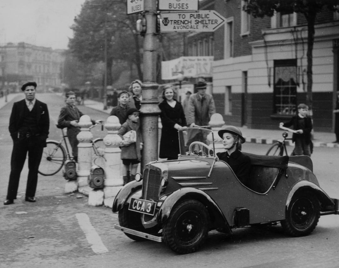 A young girl drives a small vintage car at a street intersection, while a group of adults and children, some with bicycles, watch from the sidewalk near a signpost. The scene appears to be from the mid-20th century.