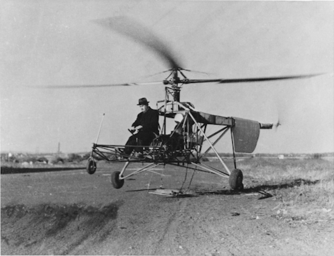 A person wearing a coat and hat pilots an early model autogyro, hovering just above a paved road in an open, grassy landscape. The rotor blades are spinning, and the machine is partially lifted off the ground.