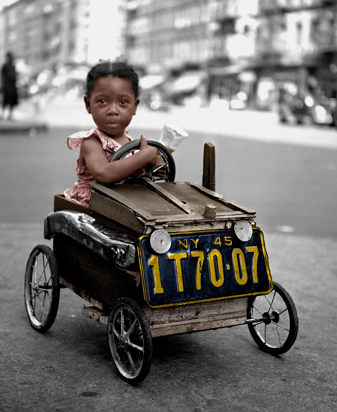 A young girl sits in a homemade toy car with a 1945 New York license plate, holding the steering wheel on a city street. The image is mostly black and white, except for the girl and the license plate, which are in color.