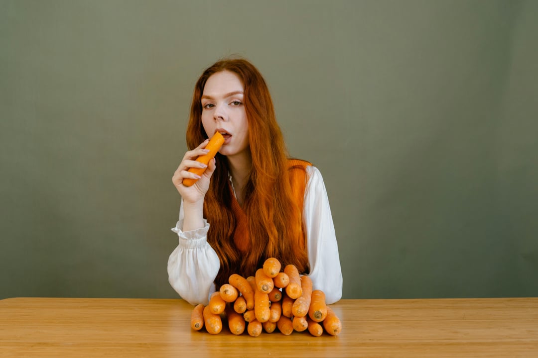 A young woman with long red hair sits at a table covered with a pile of carrots, holding and eating one carrot, against a plain green background.
