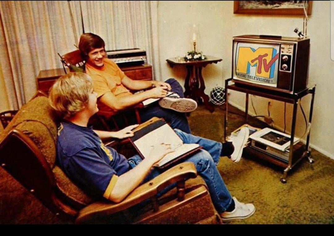 Two young men sit in a living room watching MTV on an old television set. One is seated on a couch with a notebook on his lap, both are smiling. The room is furnished with simple decor and a carpeted floor.