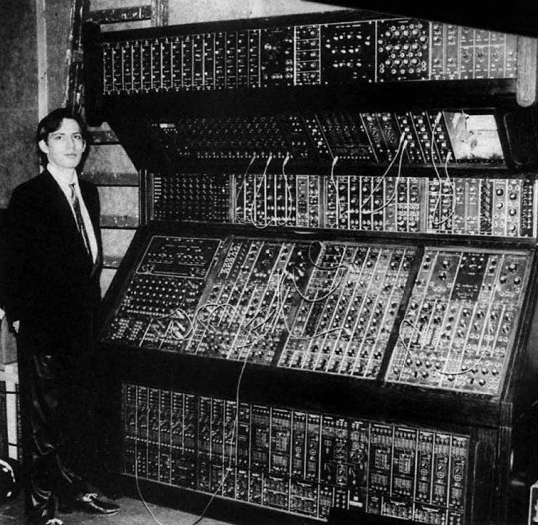 A person in a suit stands next to a large, complex modular synthesizer with numerous dials, switches, and patch cables, filling an entire wall. The image is black and white.