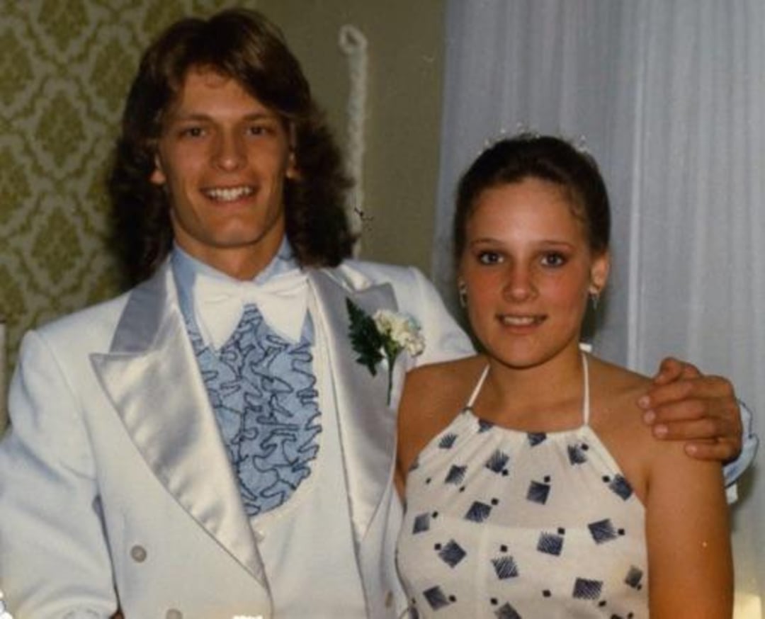 A young man in a white tuxedo with a ruffled blue shirt stands next to a young woman in a halter dress with a geometric pattern. They are smiling, posing together indoors.