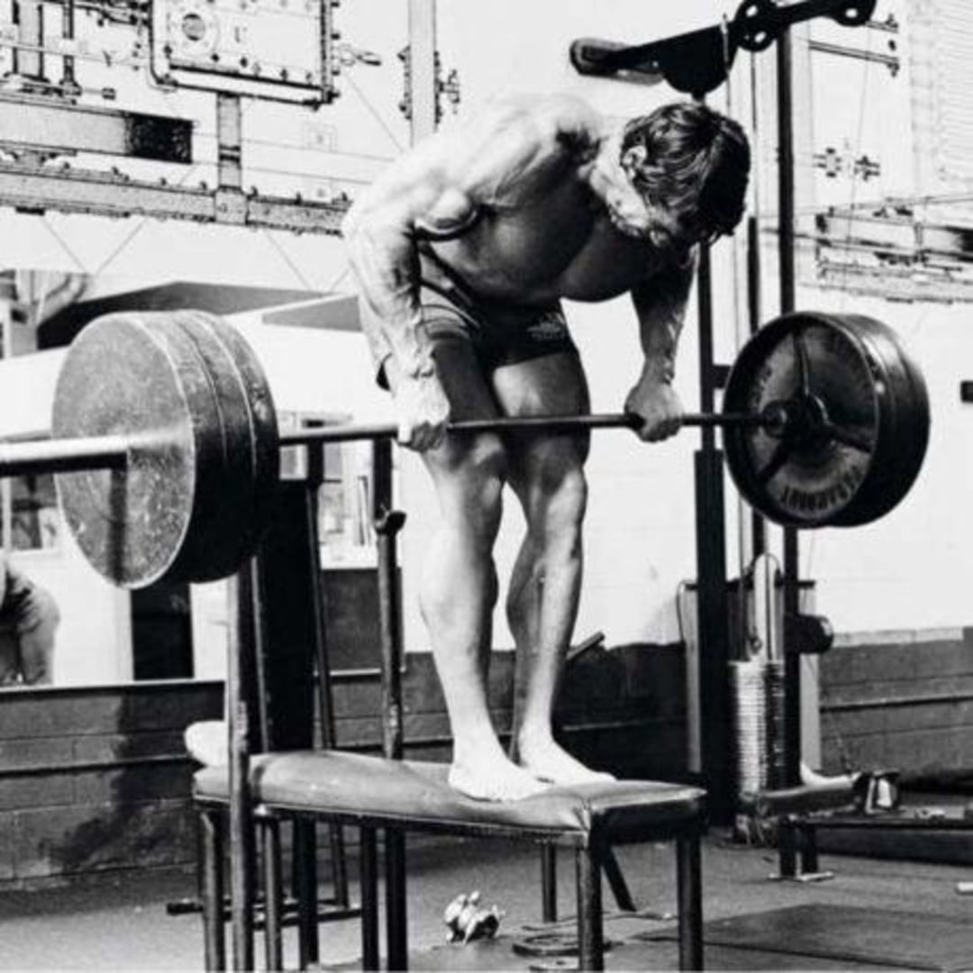 A muscular man in shorts stands barefoot on a bench, performing a bent-over barbell row with heavy weights in a gym, focusing intently on his form.