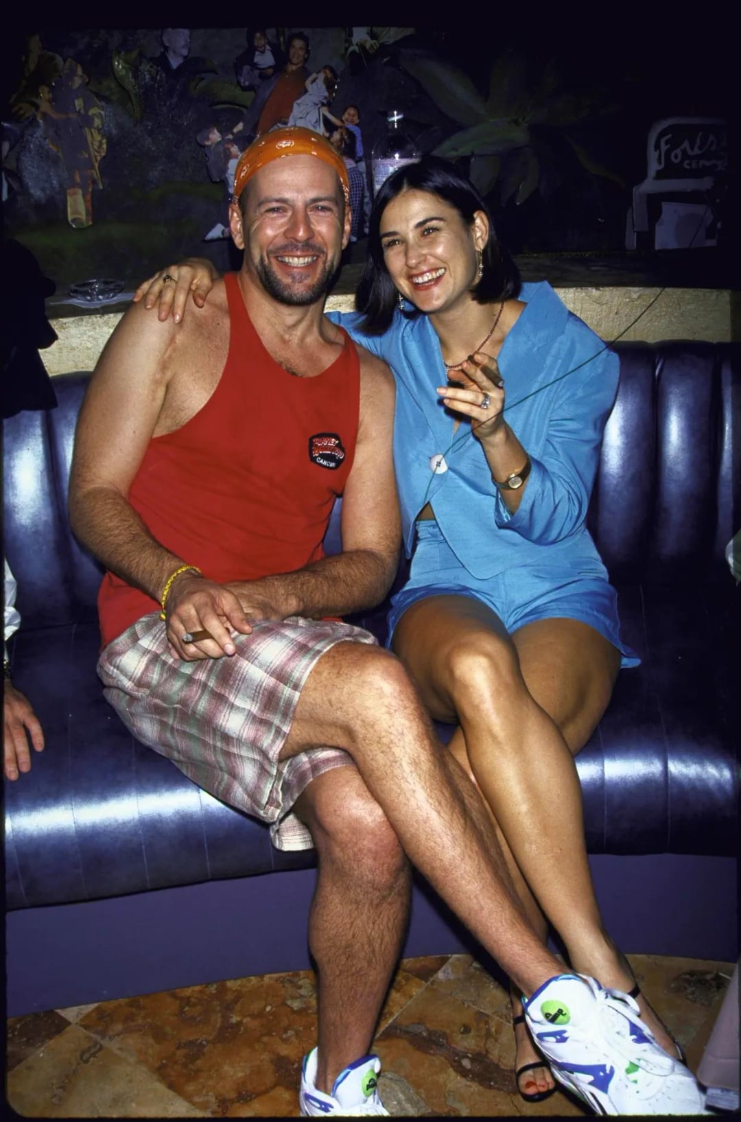 A smiling man in a red tank top, plaid shorts, and orange headband sits on a blue couch with a woman in a light blue outfit and white sneakers. They both have their arms around each other and appear cheerful.