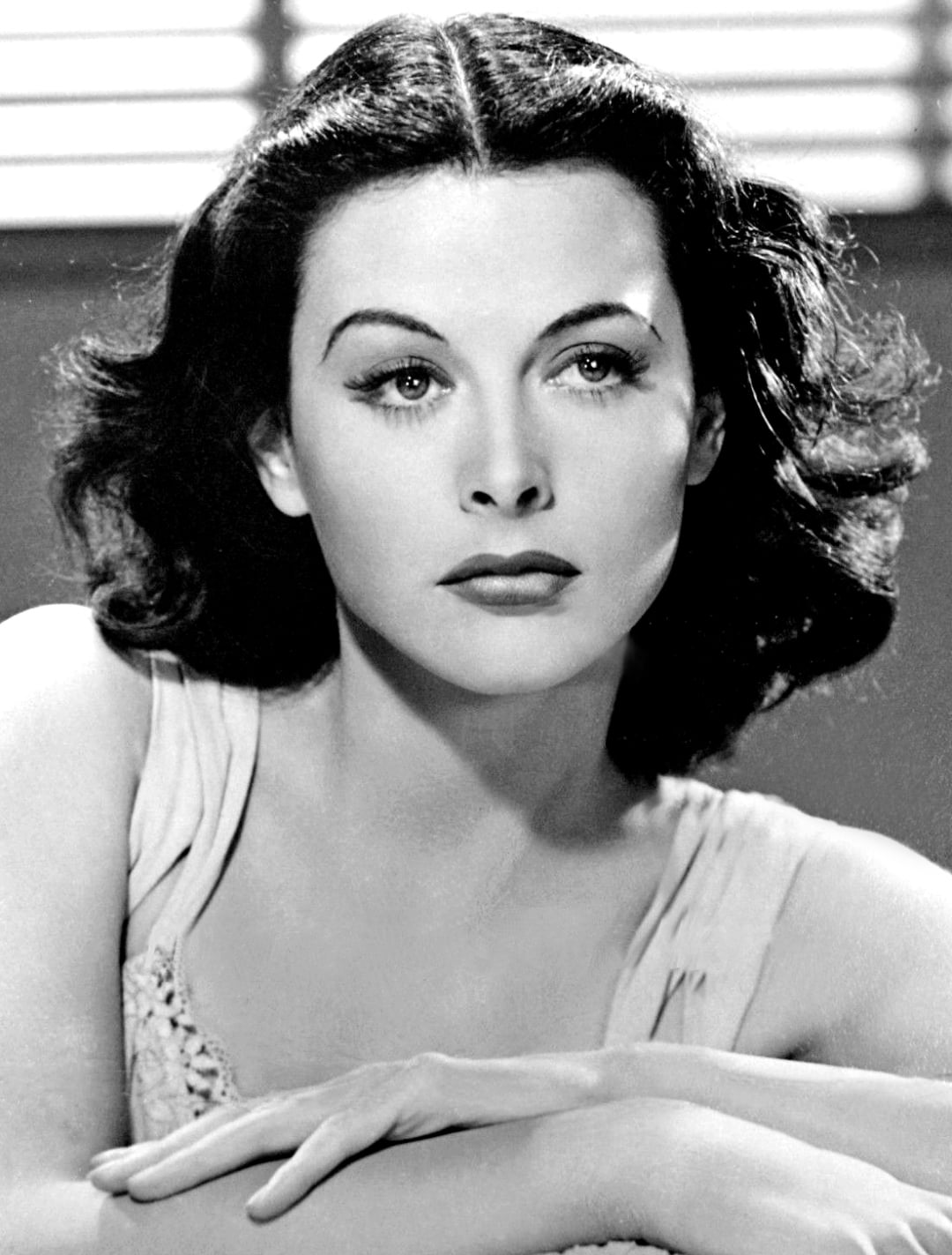 Black and white portrait of a woman with wavy dark hair, wearing a sleeveless top, gazing slightly to the side with her arms crossed in front of her, in front of a background with horizontal lines.